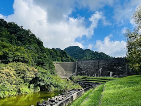 A landscape featuring a large dam set against a backdrop of lush, green forests and hills under a partly cloudy blue sky. The foreground includes a grassy path and a body of water reflecting the vegetation. The scene conveys a sense of serene natural beauty.