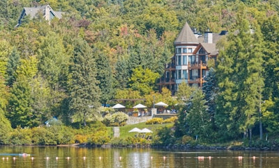A large, elegant house with a conical roof is nestled among dense green trees and foliage near a body of water. Several balconies and large windows are visible on the house. There are outdoor sitting areas with white umbrellas and tables on a landscaped terrace. The water in the foreground reflects the surrounding greenery and is dotted with buoy markers.