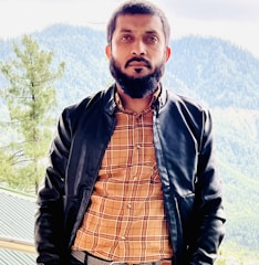 A bearded man stands outdoors wearing a black leather jacket over a plaid shirt. Behind him is a scenic background featuring green mountains and trees, suggesting a serene, natural setting.