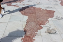A detailed stone map on the ground representing parts of the world with decorative illustrations of ships and sea creatures. People walking nearby cast shadows onto the map.