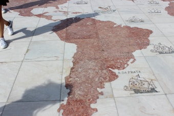 A detailed stone map on the ground representing parts of the world with decorative illustrations of ships and sea creatures. People walking nearby cast shadows onto the map.