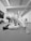 Two individuals are engaged in a jiu-jitsu practice on a mat in a sports facility. One person is in a dominant position over the other, who is on their back, both wearing traditional martial arts attire. In the background, another person is observing or possibly practicing independently. The room is well-lit and appears to be spacious with martial arts equipment along one wall.
