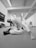 Two individuals are engaged in a jiu-jitsu practice on a mat in a sports facility. One person is in a dominant position over the other, who is on their back, both wearing traditional martial arts attire. In the background, another person is observing or possibly practicing independently. The room is well-lit and appears to be spacious with martial arts equipment along one wall.