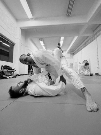 Two individuals are engaged in a jiu-jitsu practice on a mat in a sports facility. One person is in a dominant position over the other, who is on their back, both wearing traditional martial arts attire. In the background, another person is observing or possibly practicing independently. The room is well-lit and appears to be spacious with martial arts equipment along one wall.
