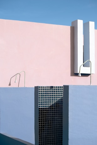 A minimalist architectural scene featuring geometric shapes and structures. The composition includes a combination of pastel pink and blue walls, along with a section of black grid-like tiles. Two thin fixtures are protruding from the walls, adding a modern touch to the overall aesthetic.
