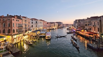 A picturesque canal flanked by historic buildings with warm lights shining from cafes and shops. Gondolas glide smoothly through the water, with people enjoying the scenic beauty. The sky displays a gentle gradient from blue to sunset hues.