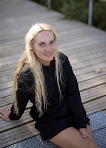 A person with long blonde hair and braces is sitting on a wooden boardwalk. The individual is wearing a black outfit and smiling while looking up at the camera. The background is slightly blurred, with hints of greenery visible.