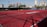 A sports court with a red playing surface and white lines, surrounded by a black mesh fence. In the background, tall buildings are visible, along with some trees and a few yellow chairs positioned near the fence.