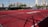 A sports court with a red playing surface and white lines, surrounded by a black mesh fence. In the background, tall buildings are visible, along with some trees and a few yellow chairs positioned near the fence.