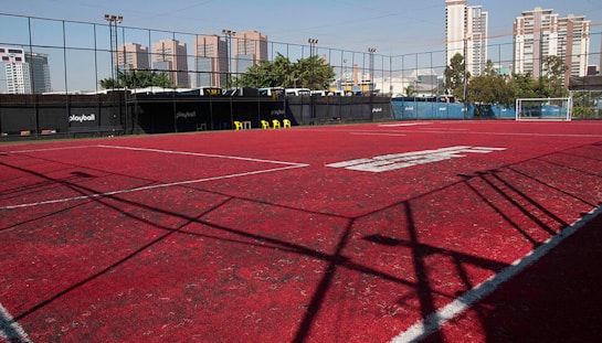 A sports court with a red playing surface and white lines, surrounded by a black mesh fence. In the background, tall buildings are visible, along with some trees and a few yellow chairs positioned near the fence.