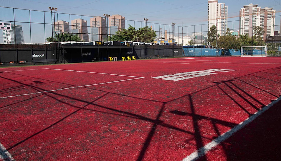 A sports court with a red playing surface and white lines, surrounded by a black mesh fence. In the background, tall buildings are visible, along with some trees and a few yellow chairs positioned near the fence.