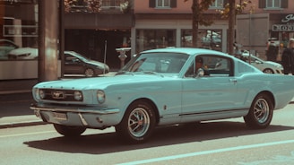A vintage light blue Ford Mustang is parked on a city street, with several modern cars and pedestrians in the background. The overall scene is bright and sunny, suggesting a pleasant day.