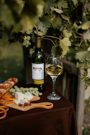 A rustic scene featuring a bottle of white wine, a filled wine glass, a cutting board with blue cheese, grapes, olives, and a baguette, all placed on a dark brown tablecloth. Grapevine leaves and grapes hang overhead, adding a natural and inviting vibe.