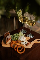 A rustic wooden cutting board holds green grapes, blue cheese, sliced bread, pretzel rings, and almonds. Two glasses of white wine are positioned nearby. The setting seems to be outdoors, with a dark, natural background.