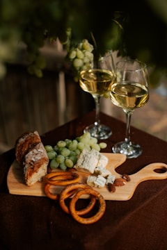 A rustic wooden cutting board holds green grapes, blue cheese, sliced bread, pretzel rings, and almonds. Two glasses of white wine are positioned nearby. The setting seems to be outdoors, with a dark, natural background.