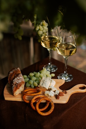 A rustic wooden cutting board holds green grapes, blue cheese, sliced bread, pretzel rings, and almonds. Two glasses of white wine are positioned nearby. The setting seems to be outdoors, with a dark, natural background.