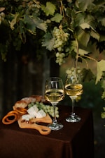 Two glasses of white wine are placed on a table covered with a brown cloth. Nearby, there is a wooden cutting board with slices of blue cheese, almonds, pretzels, and pieces of bread. Above, green grapevines with clusters of grapes hang down, adding a natural, vineyard atmosphere.
