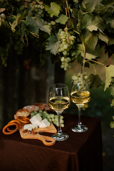 Two glasses of white wine are placed on a table covered with a brown cloth. Nearby, there is a wooden cutting board with slices of blue cheese, almonds, pretzels, and pieces of bread. Above, green grapevines with clusters of grapes hang down, adding a natural, vineyard atmosphere.