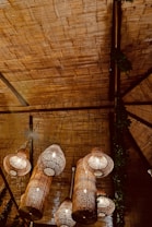 Woven rattan or bamboo pendant lights hanging from a textured ceiling, casting a warm, cozy glow. The ceiling appears to be made of a natural material, possibly wood or bamboo, with a section of green foliage adding a touch of nature.