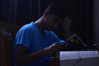 A person wearing a blue shirt is sitting at a table, reading a book. The setting appears dark, with shadows cast around the room, giving a focused and introspective mood. The individual appears to be deeply concentrated, resting their head on one hand while holding the book with the other.
