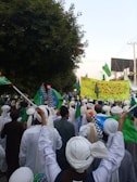 A large group of people wearing traditional clothing and turbans are participating in a public gathering or march. Many individuals are holding green flags with different designs. There is a large yellow banner with text written on it in a script language. Trees and buildings are visible in the background.
