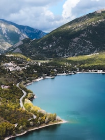 A picturesque landscape featuring a serene lake with turquoise waters surrounded by lush green hills and mountains. A winding road traces the edge of the lake, bordered by dense clusters of trees. A small boat is seen on the water, emphasizing the tranquility of the scene.