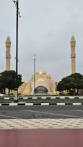 A mosque with two tall minarets flanking its main dome is set against a cloudy sky. The architecture features pale yellow tones and geometric shapes. Trees are symmetrically placed on either side, and the surrounding street is lined with streetlights and a neatly paved sidewalk.