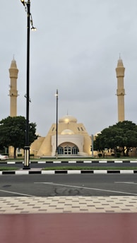 A mosque with two tall minarets flanking its main dome is set against a cloudy sky. The architecture features pale yellow tones and geometric shapes. Trees are symmetrically placed on either side, and the surrounding street is lined with streetlights and a neatly paved sidewalk.