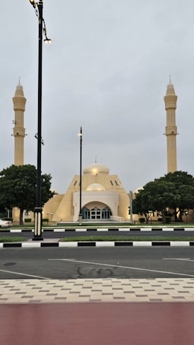 A mosque with two tall minarets flanking its main dome is set against a cloudy sky. The architecture features pale yellow tones and geometric shapes. Trees are symmetrically placed on either side, and the surrounding street is lined with streetlights and a neatly paved sidewalk.