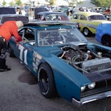 A classic car with the number 71 is displayed in a parking lot among other vintage vehicles. The car's hood is open, revealing its engine and internal parts. A person wearing a red jacket and knee brace is leaning into the car's window area, seemingly inspecting or working on the interior. Cars of various colors and styles are visible in the background, along with a crowd of people walking around.