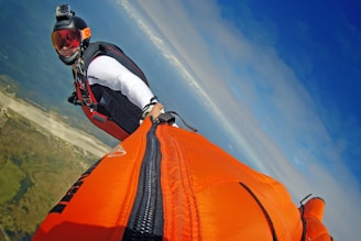 A person wearing a wingsuit is captured mid-flight against a backdrop of a clear blue sky and distant landscape below. The wingsuit is bright orange, and the person is wearing a helmet equipped with a camera. The scene conveys a sense of adventure and freedom.