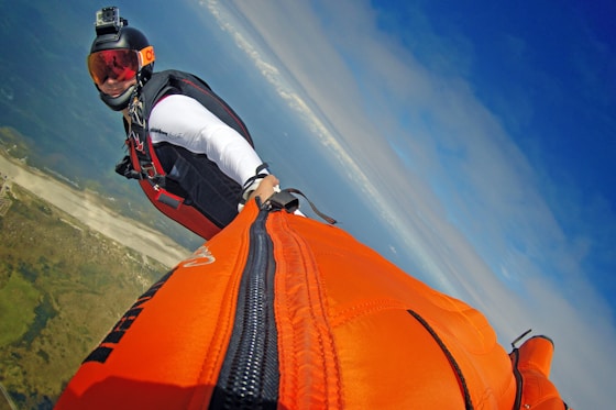 A person wearing a wingsuit is captured mid-flight against a backdrop of a clear blue sky and distant landscape below. The wingsuit is bright orange, and the person is wearing a helmet equipped with a camera. The scene conveys a sense of adventure and freedom.