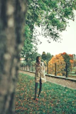 A woman in a beige trench coat walking through a sunlit park with autumn leaves around her