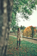 A woman in a beige trench coat walking through a sunlit park with autumn leaves around her
