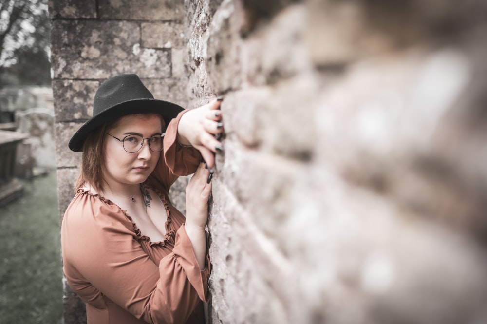a woman wearing glasses and a hat leaning against a brick wall