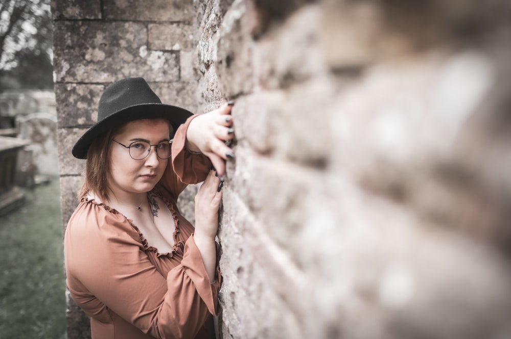 Una mujer con gafas y un sombrero apoyada contra una pared de ladrillos