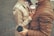 A couple stands closely together facing each other, each holding a cup of coffee with lids. They are wearing brown jackets, and the ground is paved with bricks.