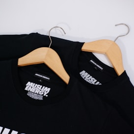 Two black T-shirts with the words 'Muslim Energy' printed in bold white letters are displayed on wooden hangers. The hangers have silver metal hooks and are placed against a white background.