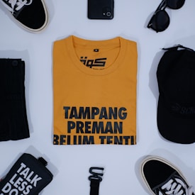 A neatly arranged flat lay featuring a mustard yellow t-shirt with bold black lettering at the center. Surrounding the shirt are various items: a pair of black sneakers with white laces, a black smartphone, round black sunglasses, a black cap, a black pouch with 'TALK LESS DO' printed in white, and a black belt.