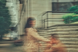 A blurred image capturing a moment between two people on a city sidewalk. One person is standing, while the other appears to be kneeling and holding the hand of the standing person. They are next to a brick wall and there is greenery nearby. The scene gives a sense of motion and urgency.