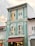 A three-story heritage building with pastel green walls and white details. It features tall, narrow windows with wooden shutters, some of which are open. The building has a brown awning on the ground floor, under which there are three hanging lights. The adjacent buildings have similar traditional architectural styles, with one in white.