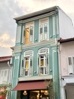 A three-story heritage building with pastel green walls and white details. It features tall, narrow windows with wooden shutters, some of which are open. The building has a brown awning on the ground floor, under which there are three hanging lights. The adjacent buildings have similar traditional architectural styles, with one in white.