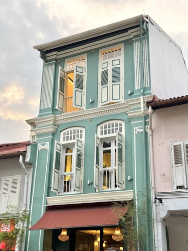 A three-story heritage building with pastel green walls and white details. It features tall, narrow windows with wooden shutters, some of which are open. The building has a brown awning on the ground floor, under which there are three hanging lights. The adjacent buildings have similar traditional architectural styles, with one in white.