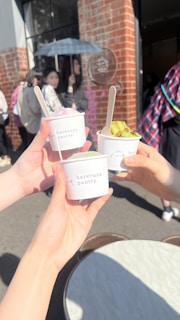 Three people holding small cups of ice cream or dessert with wooden spoons, labeled 'Hareruya Pantry'. The setting appears to be outside a store or café, with a brick wall and a glass window in the background. There are several people in the background, one holding a blue umbrella.