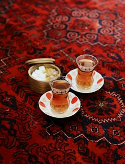 Two glasses of tea on white saucers rest atop a richly patterned red and dark blue carpet. A small wooden container filled with sugar cubes and rose petals is situated next to the tea glasses.