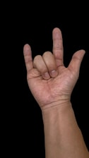 A hand gestures in a 'rock on' or 'I love you' sign, with the thumb, index finger, and pinky extended. The background is black, highlighting the skin tone and texture.