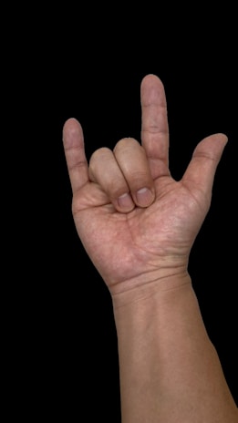 A hand gestures in a 'rock on' or 'I love you' sign, with the thumb, index finger, and pinky extended. The background is black, highlighting the skin tone and texture.