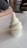 A creamy, soft-serve ice cream swirl sits neatly in a white paper cup atop a smooth, white surface. Nearby, a person's arm rests on the table, and there are blurred background elements, including orange chairs and a window.