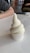 A creamy, soft-serve ice cream swirl sits neatly in a white paper cup atop a smooth, white surface. Nearby, a person's arm rests on the table, and there are blurred background elements, including orange chairs and a window.