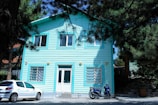 A turquoise-painted wooden house with two floors is surrounded by tall pine trees. The house has white-framed windows and a front door with a small set of stairs leading up to it. In front of the house, a white car and a blue scooter are parked on a paved area.